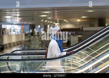 Afroamerikanischer Passagiermann mit Koffer steht auf Rolltreppe, hält Handlauf im Flughafenterminal Stockfoto