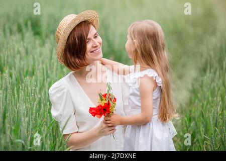 Mutter und Tochter gehen auf dem Feld. Ein blondes Mädchen mit langen Haaren gibt ihrer Mutter einen Strauß Mohnblumen für den Muttertag. Stockfoto