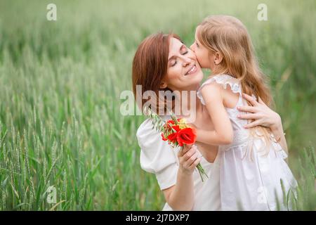 Mutter und Tochter gehen auf dem Feld. Ein blondes Mädchen mit langen Haaren gibt ihrer Mutter einen Strauß Mohnblumen für den Muttertag. Stockfoto