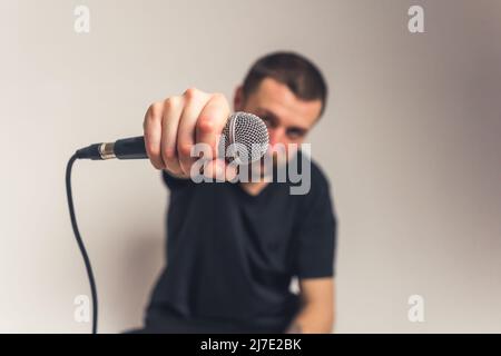 Kurzhaariger kaukasischer bärtiger Mann, der das Mikrofon in der Hand an die Kamera hält, Vordergrundfokus. Studio auf grauem Hintergrund. Hochwertige Fotos Stockfoto
