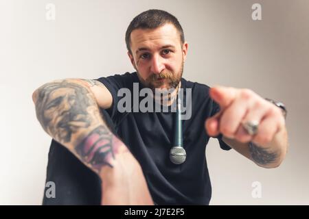 Ernster wütender kaukasischer bärtiger Mann mit Tattoo-Ärmeln und einem Mikrofon über seiner Schulter, das auf die Kamera auf grauem Hintergrund zeigt. Hochwertige Fotos Stockfoto