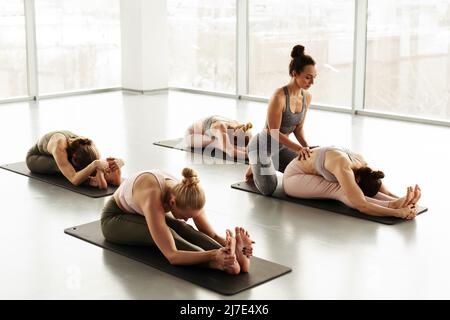 Junge Yoga-Trainerin schiebt die untere Rückseite der Schülerin, während sie ihre Asana beim Yoga-Kurs anpasst Stockfoto