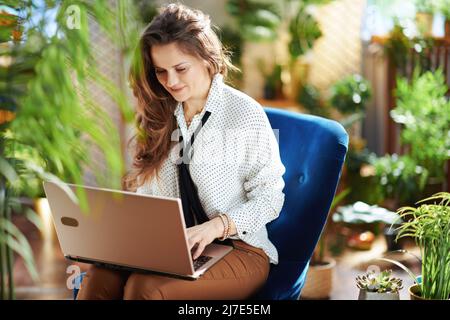 Green Home. Lächelnd stilvolle 40 Jahre alte Hausfrau mit langen welligen Haaren mit Laptop in beige Hosen und Bluse sitzt in einem blauen Sessel in der modernen Stockfoto