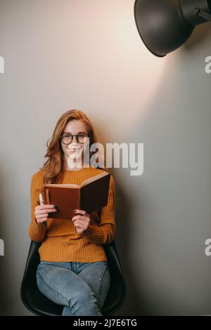 Porträt einer kaukasischen rothaarigen Frau, die ein Notizbuch hält und auf einem Stuhl unter dem Scheinwerfer sitzt, die Kamera anschaut und lächelt. Hochformat ein Stockfoto