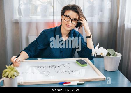Der Lehrer posiert auf dem Schreibtisch und sieht sich die Kamera an, die Sätze auf einem weißen Brett mit einem blauen Marker schreibt. Sie besucht zusammen mit ihr ein virtuelles Klassenzimmer Stockfoto