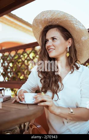 Lächelnde trendige 40-jährige Hausfrau in weißem Hemd mit einer Tasse Kaffee und einem Hut, die auf der Terrasse des Gästehaushotels am Tisch sitzt. Stockfoto