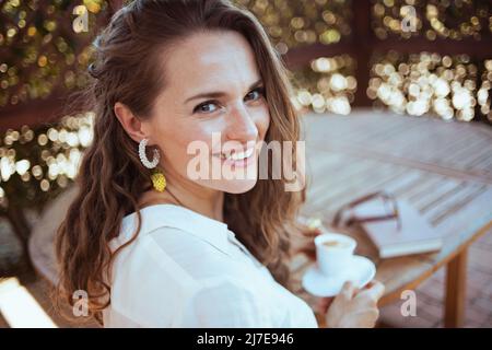 Porträt einer glücklichen jungen Frau im weißen Hemd mit einer Tasse Kaffee, einem Buch und einer Brille, die auf der Terrasse am Tisch sitzt. Stockfoto