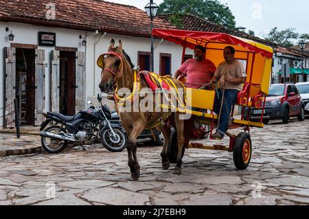 Ein farbenfroher Wagen, der von einem braunen Pferd gezogen wird, mit einem Passagier, der durch die gepflasterte Ministro Gabriel Passos Straße im historischen Zentrum von Tiradentes reitet. Stockfoto