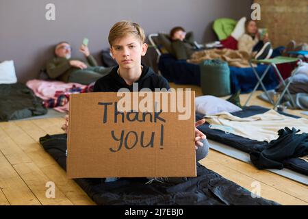 Dankbarer Jugendlicher in Casualwear, der ein Pappposter in der Hand hält, während er auf dem Boden eines geräumigen Zimmers sitzt und als vorübergehender Schutz dient Stockfoto
