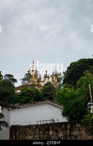 Teilansicht der Santo Antonio Mutter Kirche Türme hinter grüner Vegetation sah von Rosario Platz in Tiradentes historischen Zentrum unter bewölktem Himmel. Stockfoto