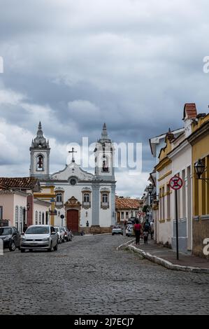 Getulio Vargas gepflasterte Straße mit Kolonialhäusern auf beiden Seiten und die große Kirche Nossa Senhora do Rosario auf der Rückseite unter bewölktem Himmel. Stockfoto