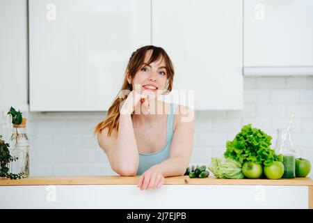 Porträt einer jungen Frau, die sich auf einem hohen Beistelltisch neben einem Haufen grünen Gemüses und Obst auf dem Tisch lehnt. In einer großen, geräumigen Küche. Tragen von BL Stockfoto