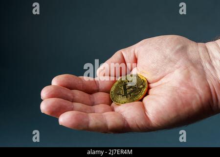goldene Bitcoin-Münze in männlicher Handfläche, grauer Hintergrund aus der Nähe Stockfoto