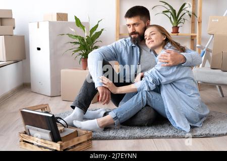 Glückliches junges Paar Mann und Frau sitzen auf dem Boden in lässiger Kleidung und umarmt. Bewegt Kartons und Möbel während eines Umzugses zu einem neuen Stockfoto