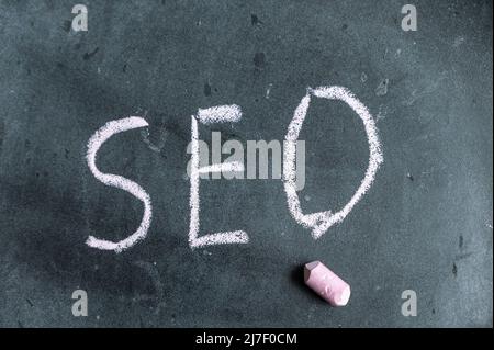 SEO. Ein Wort, das in rosa Kreide auf einer schwarzen Tafel geschrieben ist. Handgeschriebener Text. Daneben hängt ein Stück farbige Kreide. Stockfoto