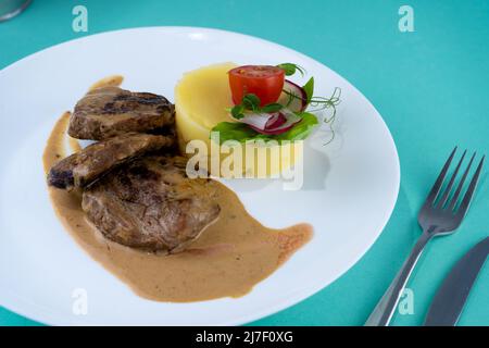 Fleischbällchen mit Kartoffelpüree auf einem weißen Teller. Fleisch in saurer Rahmsoße. Tomaten und Basilikum. Essen im Restaurant. Mittagspause. Medaillons. Stockfoto