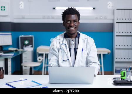 Lächelnder afroamerikanischer Klinikspezialist, der am Schreibtisch sitzt, während er einen Laptop benutzt. Der Krankenhausarzt mit weißer Uniform sitzt am Schreibtisch im Arbeitsbereich und verwendet dabei einen modernen Computer Stockfoto