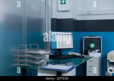 Leere Arztpraxis mit niemand darin, ausgestattet mit medizinischen Geräten, Arbeitscomputer auf dem Schreibtisch und erste-Hilfe-Kasten an der Wand. Moderner Krankenhausraum mit Rollboxen und Zwischenablageberichten. Stockfoto