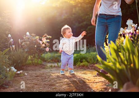 Ein süßes kleines Kind schaut seine Mutter an und greift zu ihr. Im Hintergrund ein Sommergarten und blühende Sträucher. Alles gute zum Kindertag. Stockfoto