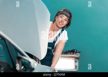 Ein Porträt einer jungen lächelnden Mechanikerin in Gläsern und Overalls blickt hinter der Motorhaube eines Autos hervor. Das Konzept der Frauenarbeit in der männlichen PR Stockfoto