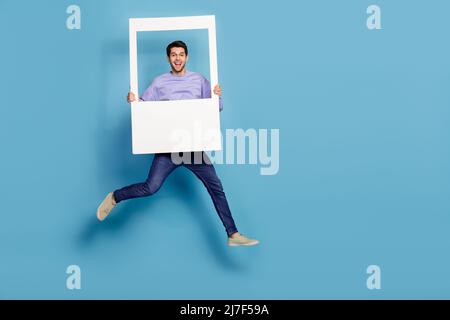 In voller Länge Körpergröße Ansicht der attraktiven fröhlichen Kerl springen halten Fotorahmen Fenster isoliert über hellen blauen Hintergrund Stockfoto