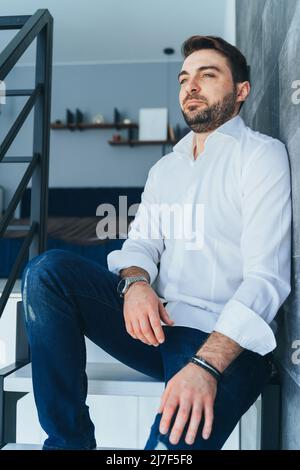 Vertikales Bild von gutaussehenden unrasierten Mann, Geschäftsmann in der Mode und trendige formelle Kleidung, wegschauen. Drinnen auf Treppen sitzen. Sich entspannt fühlen Stockfoto