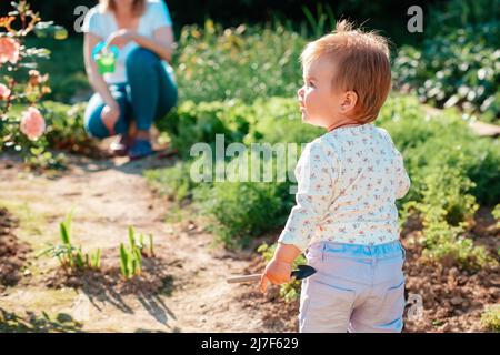 Gartenarbeit. Das niedliche Kind steht im Garten mit einer kleinen Hacke in der Hand. Rückansicht. Im Hintergrund sitzt eine Frau neben einem Blumenbett mit pla Stockfoto