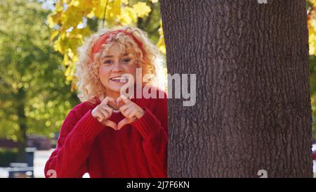 Lächelndes blondes kaukasisches Mädchen zeigt Herzzeichen, das am Baum im Park steht. Hochwertige Fotos Stockfoto