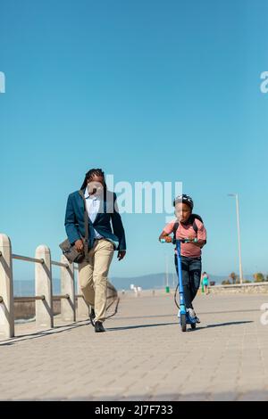 afroamerikanischer Junge, der auf dem Push-Roller des Vaters auf der Promenade gegen den klaren blauen Himmel unterwegs ist Stockfoto