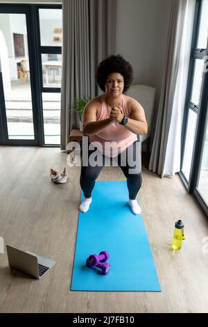 Porträt einer afroamerikanischen Frau mit mittlerem Erwachsenen, die Kniebeugen macht, während sie zu Hause auf einer Übungsmatte trainiert Stockfoto