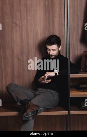 Eleganter Mann sitzt mit einem Telefon in einem Loft-Innenraum. Bärtiger Kerl nutzt Smartphone für die Kommunikation und soziale Netzwerke Stockfoto