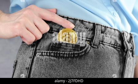 Goldene Bitcoin-Münze in einer weiblichen Nahaufnahme. Eine Frauenhand steckt die goldene Bitcoin in ihre Jeanstasche. Kryptowährung. Stockfoto