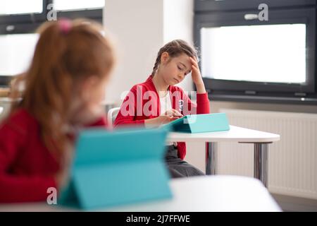 Gelangweilte Schülerin mit digitalen Tablet während des Unterrichts im Klassenzimmer an der Grundschule. Stockfoto