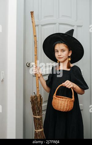 Kleines Mädchen in einem Hexenkostüm posiert vor der Tür, macht Trick oder behandelt. Einen Broomstick halten und einen Korb für Süßigkeiten halten. Stockfoto