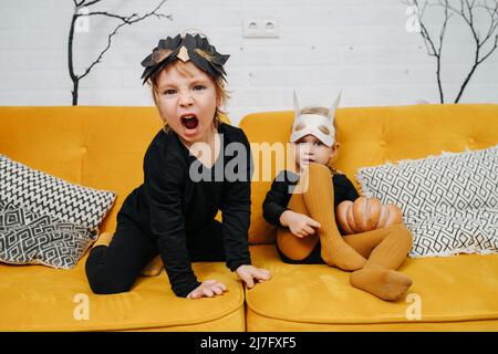 Verspielte umständliche Kinder im Vorschulalter, die zu halloween auf einer gelben Couch gekleidet waren und Masken trugen. Junge brüllt wie ein Löwe. Das Zimmer war mit leafless eingerichtet Stockfoto