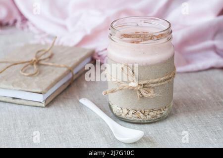 Haferflocken, gewürzt mit Joghurt, bestreut mit Zimt in einem Glas. Als nächstes gibt es ein Bastelheft und eine rosa Serviette. Stockfoto