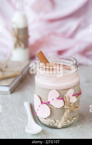 Haferflocken, gewürzt mit Joghurt, bestreut mit Zimt in einem Glas. Als nächstes kommt eine Flasche Milch. Als nächstes gibt es ein Bastelheft und eine rosa Serviette. Stockfoto