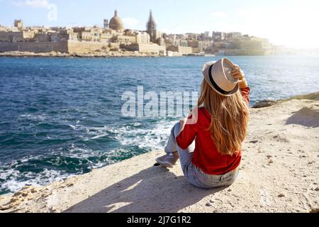 Schöne junge Frau mit Hut sitzt auf Stein am Meer mit herrlichem Panoramablick auf Valletta Stadt in Malta Stockfoto