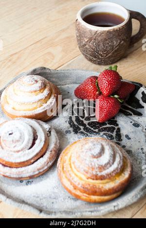 Köstliche Brötchen werden mit Zuckerpulver bestreut und rote, saftige Erdbeeren liegen auf einem hellen, handgefertigten Teller auf hellem Holzhintergrund. Als nächstes ist eine Tasse o Stockfoto