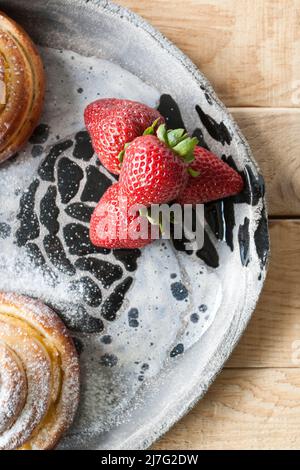 Köstliche Brötchen werden mit Zuckerpulver bestreut und rote, saftige Erdbeeren liegen auf einem hellen, handgefertigten Teller auf hellem Holzhintergrund. Stockfoto