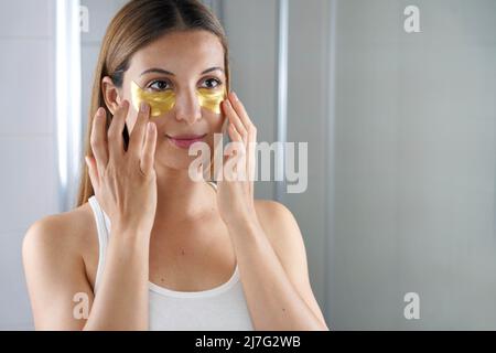 Beauty-Frau, die goldene Anti-Aging-Maske unter den Augen anwendet und sich in den Spiegel im Badezimmer sieht. Hautpflege Mädchen berühren Flecken von Stoff Maske unde Stockfoto