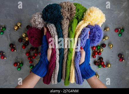 Hände von jungen schönen Frau in blauen Fäustlingen mit vielen farbigen hellen warmen Wintermützen. Auf einem grauen Hintergrund sind helle Weihnachtsdekorationen. Stockfoto