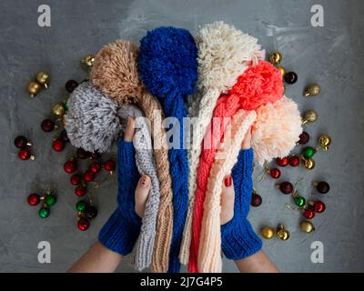 Hände von jungen schönen Frau in blauen Fäustlingen mit vielen farbigen hellen warmen Wintermützen. Auf einem grauen Hintergrund sind helle Weihnachtsdekorationen. Stockfoto