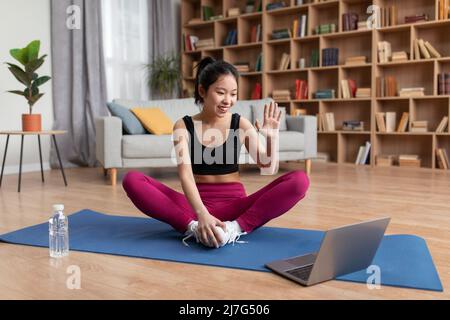 Positive asiatische Dame winkt zu Laptop-Webcam machen Videokonferenz mit Personal Trainer, sitzen auf Yoga-Matte Stockfoto