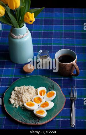 Ein gesundes und nahrhaftes Frühstück: Eine Tasse Tee; ein Teller mit Hartkoildeiern mit Kuskus. Ein Tulpenstrauß in einer Vase, eine karierte Tischdecke auf einem Schatten Stockfoto