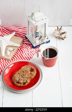 Haferflockenkekse mit Preiselbeeren liegen auf einem roten Teller, neben einer Tasse schwarzen Kaffee, einer Taschenlampe, Muscheln und einem alten Notizbuch. Die Komposition ist eingeschaltet Stockfoto