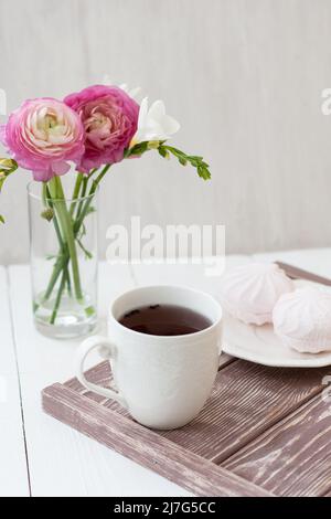 Frühling, Sommer Stillleben Szene. Eine Tasse Tee in einer weißen Tasse. Als nächstes kommt ein Bouquet mit weißer Freesie und rosa Ronunculus. Als nächstes gibt es eine weiße Platte Stockfoto