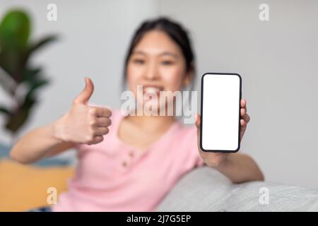 Junge asiatische Dame, die zu Hause auf der Couch sitzt, zeigt das Smartphone mit weißem leeren Bildschirm und Daumen nach oben, Nachbau Stockfoto