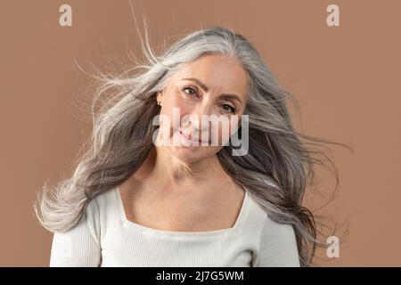 Porträt einer lächelnden älteren Frau, die mit langen und gesunden grauen, fliegenden Haaren auf braunem Studiohintergrund posiert Stockfoto