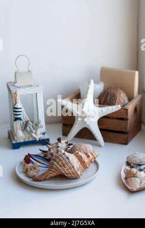 Weißer und blauer Kerzenständer in Form einer Taschenlampe. In der Nähe befindet sich ein Teller mit Muscheln und eine Holzkiste mit Kokosnuss und weißen Seesternen. Stockfoto